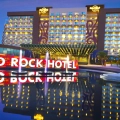 Nighttime exterior of Hard Rock Hotel Cancun's main entrance with illuminated golden guitar logos on two modern high-rise towers, red "HARD ROCK HOTEL" signage reflected in a central fountain pool, palm trees, and walkways under purple twilight skies.
