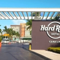 Grand entrance gate to Hard Rock Hotel Cancun under a white pergola with wooden beams, featuring a large brown sign with white "HARD ROCK HOTEL CANCUN" lettering and circular logo, flanked by palm trees, manicured shrubs, and a paved driveway leading to orange-accented resort buildings under a clear blue sky.