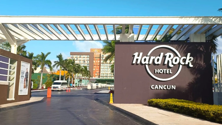 Grand entrance gate to Hard Rock Hotel Cancun under a white pergola with wooden beams, featuring a large brown sign with white "HARD ROCK HOTEL CANCUN" lettering and circular logo, flanked by palm trees, manicured shrubs, and a paved driveway leading to orange-accented resort buildings under a clear blue sky.