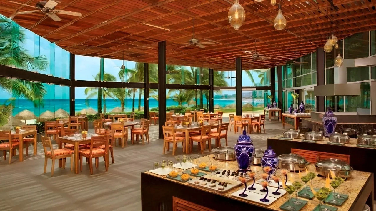 Open-air beachfront restaurant at Krystal Grand Cancun with wooden ceiling fans and hanging lanterns, rows of wooden tables with orange-cushioned chairs, a buffet station displaying covered dishes, fruits, and garnishes on a dark counter, potted plants and blue vases as decor, and expansive glass walls overlooking palm trees, white sand, and the turquoise sea.