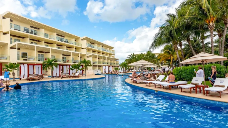 Aerial view of Azul Beach Resort Riviera Cancun showcasing multiple interconnected blue swimming pools with thatched-roof cabanas, white lounge chairs, multi-story resort buildings with balconies, palm trees, and tropical landscaping under a partly cloudy sky.
