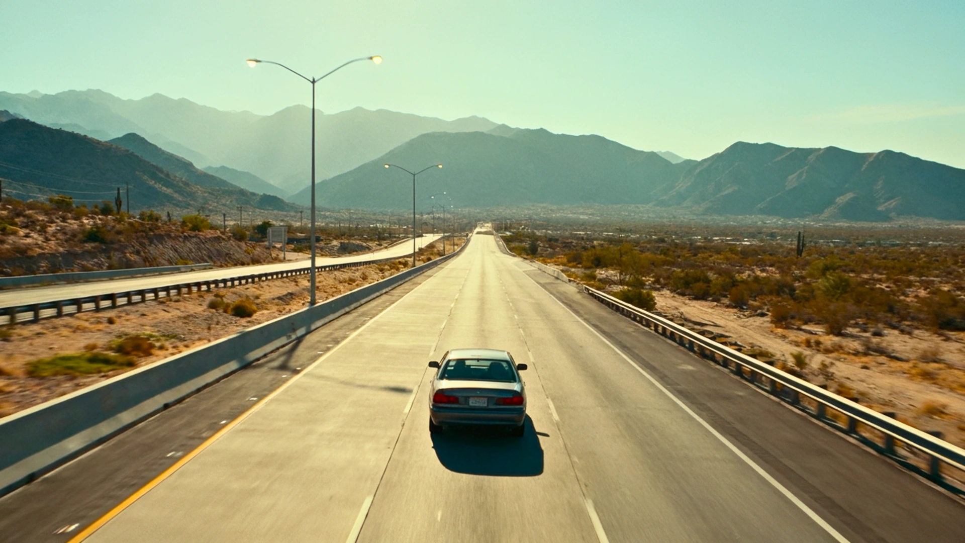 A sedan drives down a long, straight highway through a dry desert mountain valley under bright sunlight, symbolizing the start of an epic road trip.
