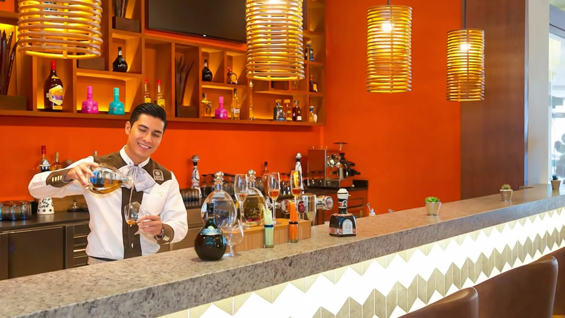 A bartender pouring a drink at a stylish bar at Hyatt Ziva Cancun, with orange walls, hanging lanterns, and an array of bottles and glassware on display.