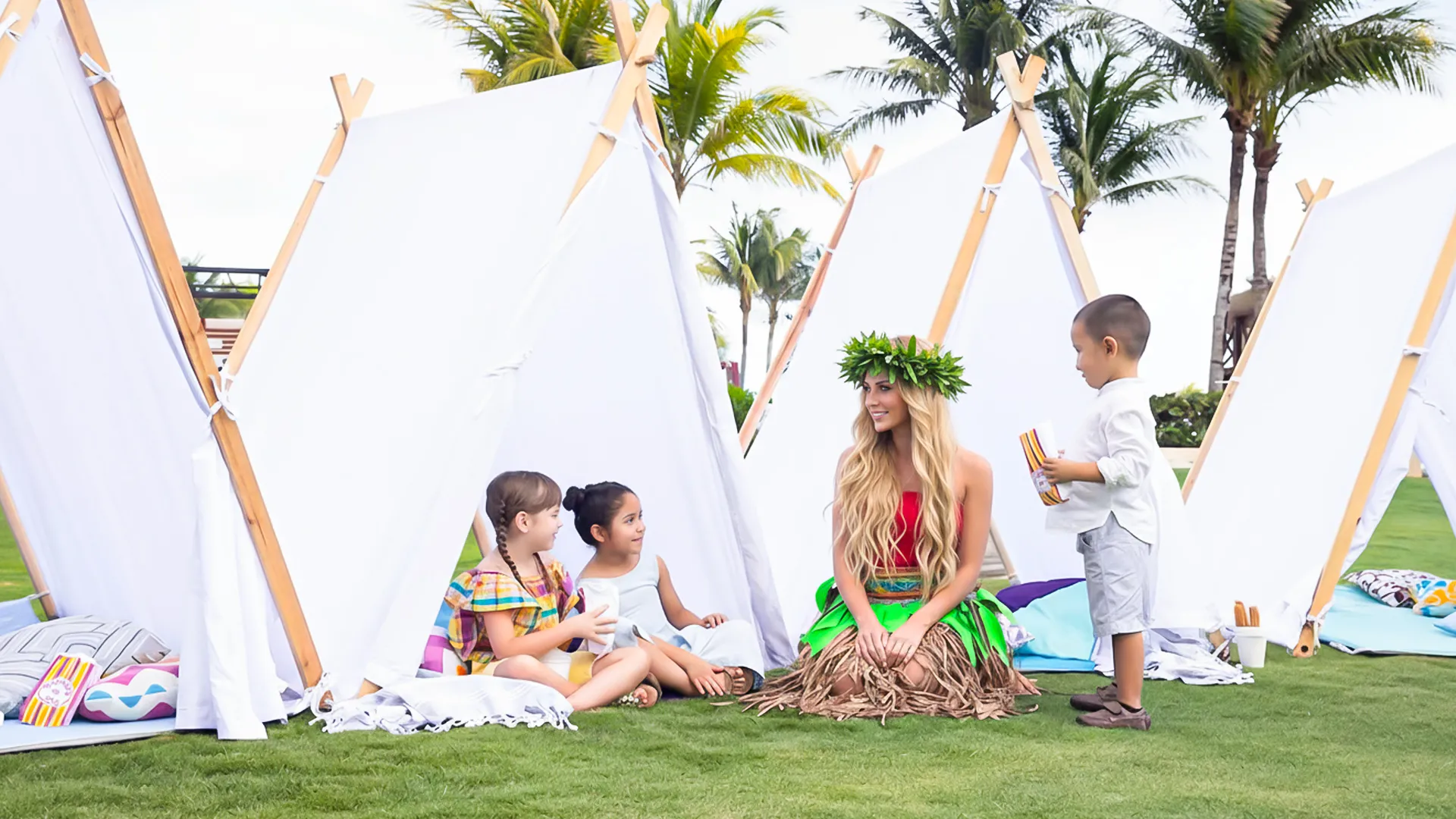 A group of children and an adult in a grass area at Hyatt Ziva Cancun, surrounded by white teepees, palm trees, and colorful cushions under a clear sky.